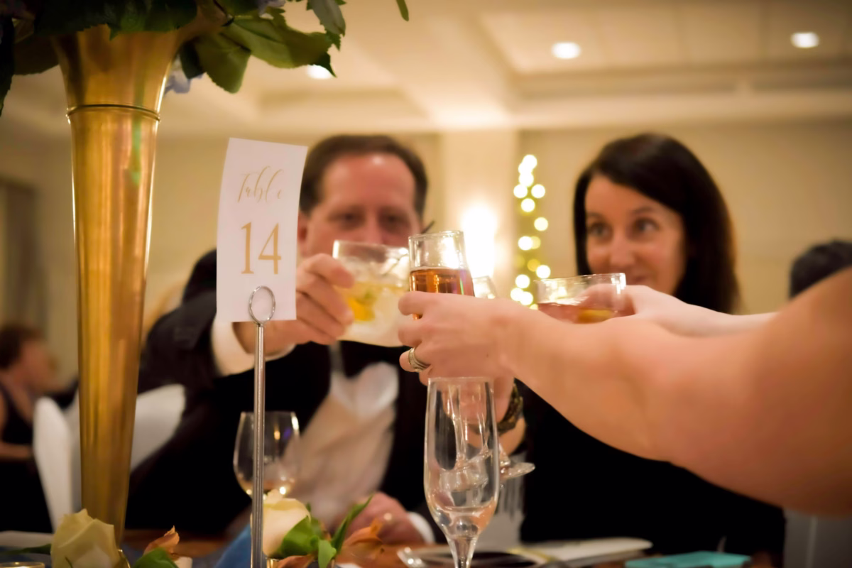 corporate awards gala champagne toast table 14 raleigh - Audio Guests raising glasses in a champagne and cocktail toast at Table 14 during a corporate awards banquet or gala event, with elegant gold floral centerpiece and holiday decor in background