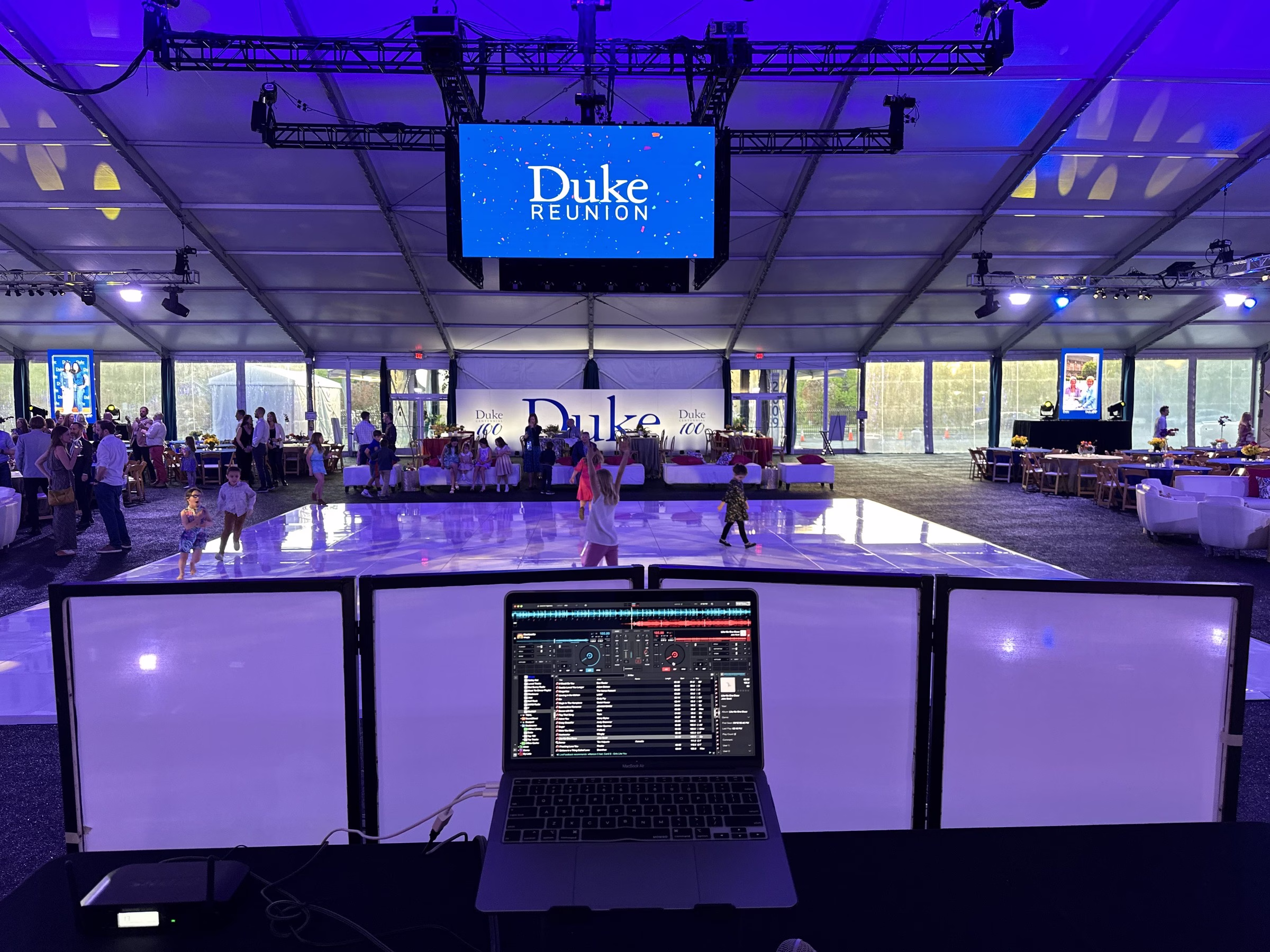 duke university reunion gala dj setup raleigh tent venue - About DJ booth with laptop running DJ software and LED facade panels overlooking a white dance floor at a Duke University Reunion corporate gala event inside a large illuminated tent venue in the Raleigh Durham Triangle area, produced by Unstoppable Events