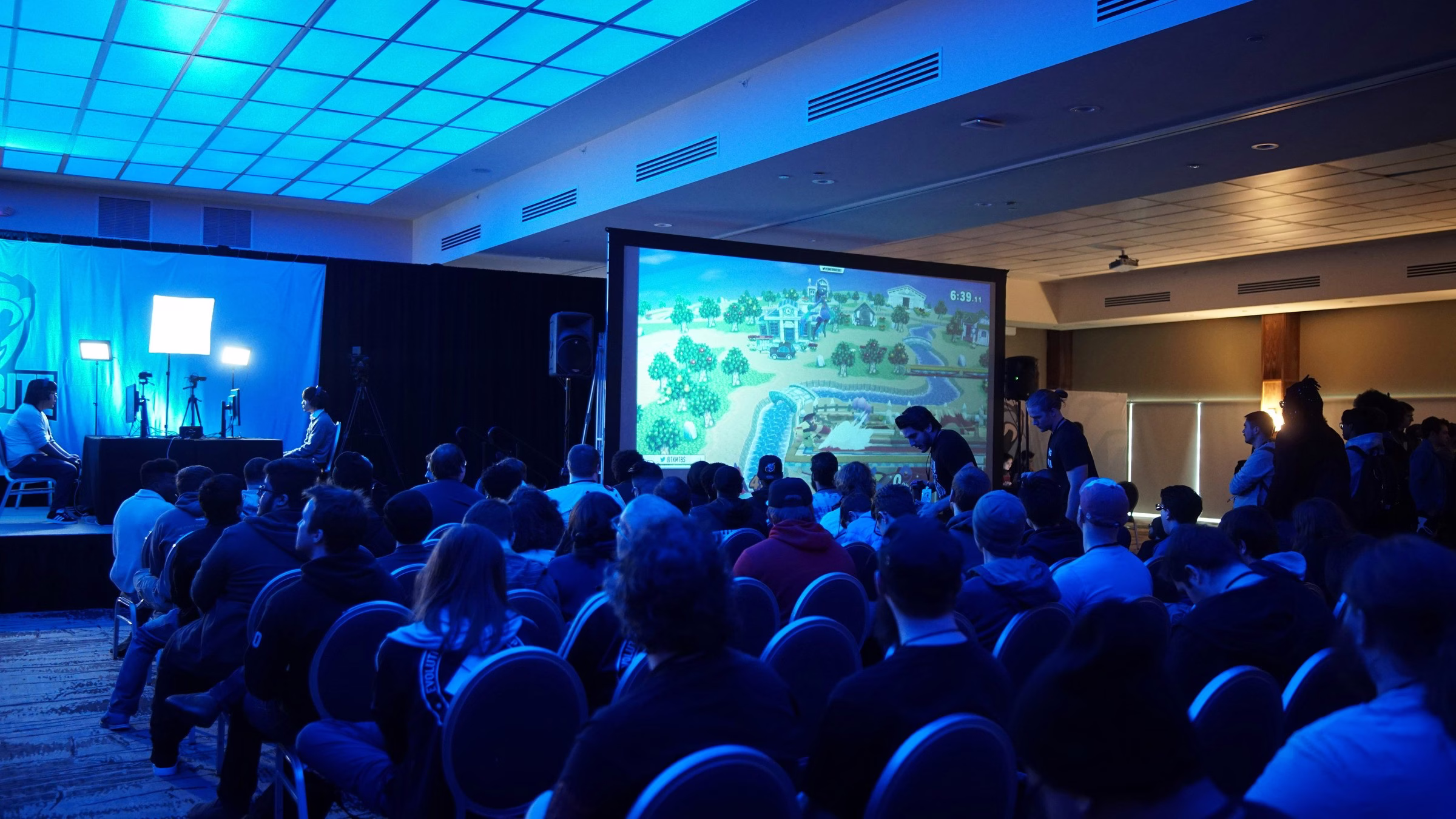 esports gaming tournament projection screen audience conference hall - Video Screens Large audience watching live esports gaming tournament on projection screen in blue-lit conference hall ballroom, with commentators on stage and PA speakers