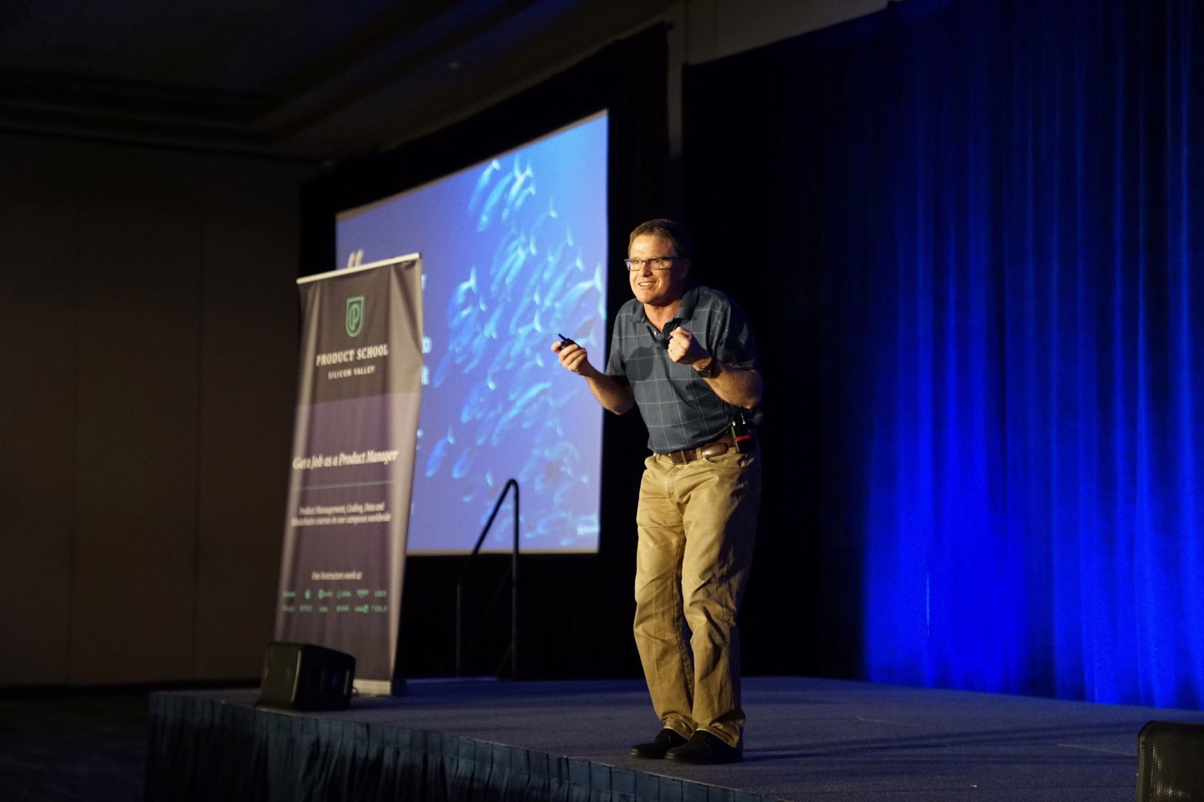 Male keynote speaker presenting on stage at corporate conference event with projection screen backdrop and Product School Silicon Valley pull-up banner, blue uplighting and pipe drape background