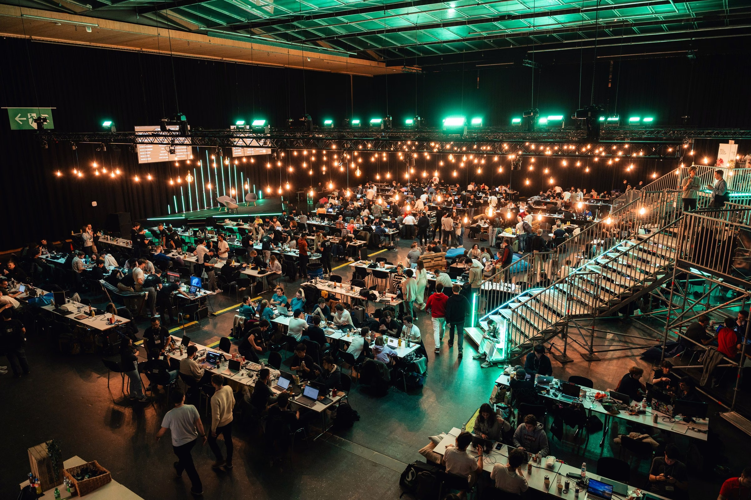 large scale hackathon corporate event production raleigh - Audio Large-scale hackathon or technology conference event production with Edison bulb string lighting design, long conference tables with laptops, stage trussing with LED uplighting, and full-room general session layout